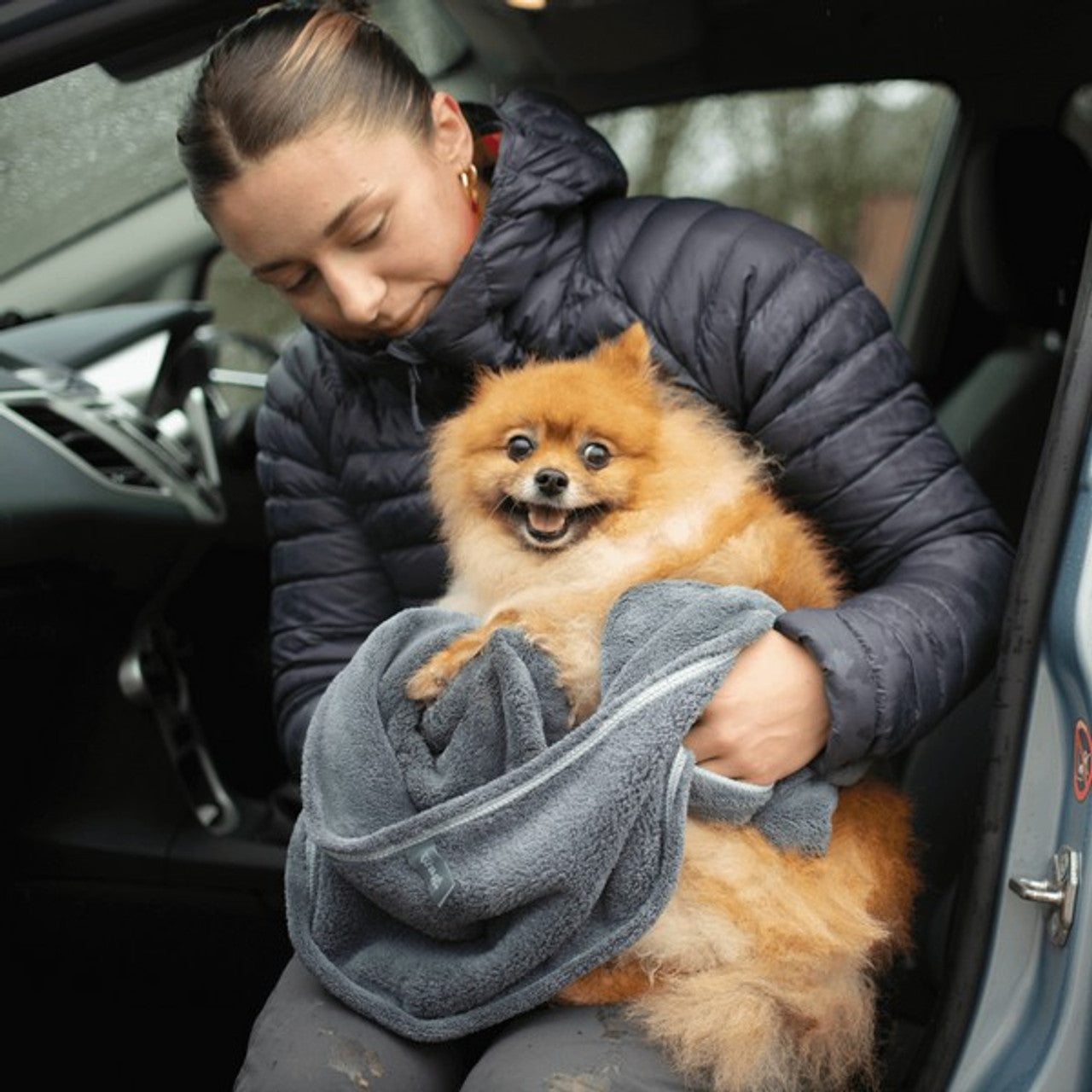 Lady drying their dog using the Henry Wag Glove Towel. Lady is sat in a car, with the dog on her lap.
