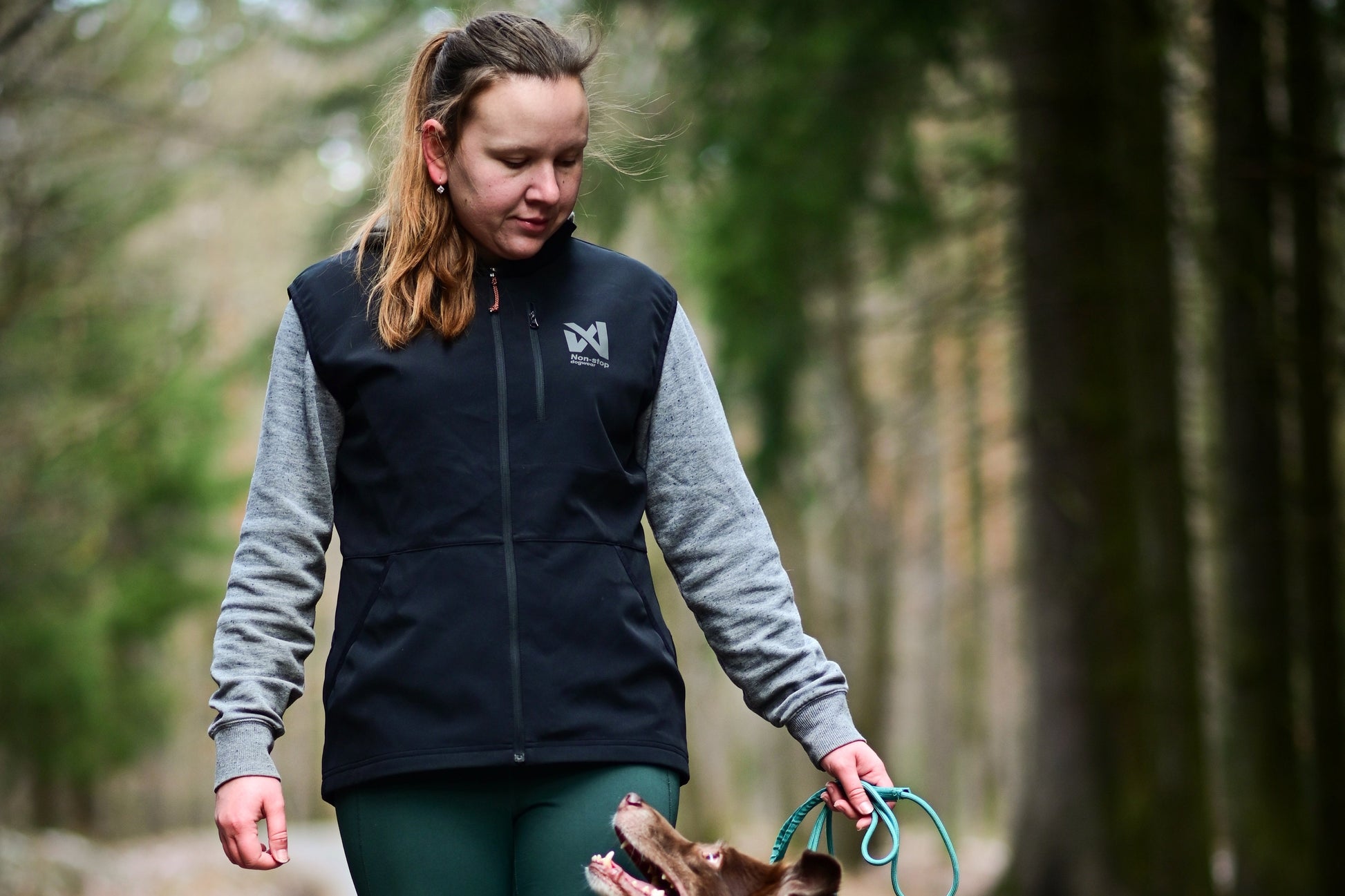 Woman walking a dog on a path in a forest, holding a dog lead while her dog looks up at her.