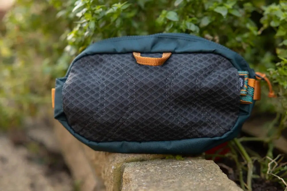 Lifestyle image of the Rachel Pohl Trail Quest Storage Pack, on a brick wall with a leafy backdrop.