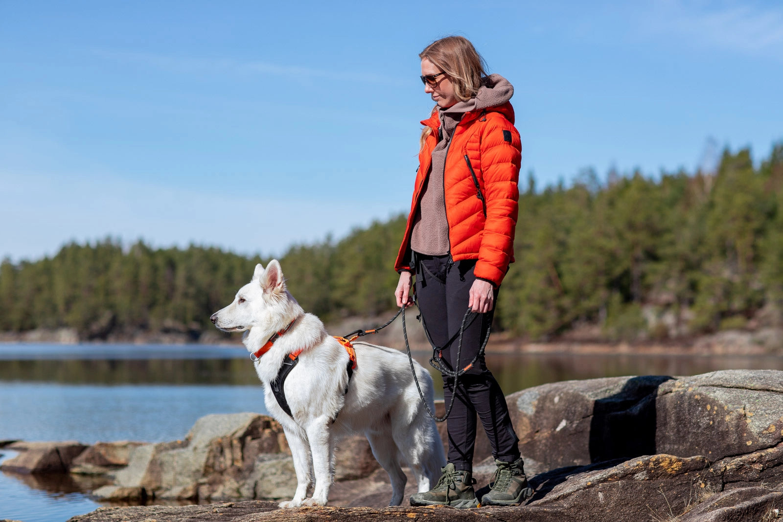 Lifestyle image of the Rope Lead, showing a white dog wearing a harness and a lady wearing an orange coat, standing on a rock with a river background.