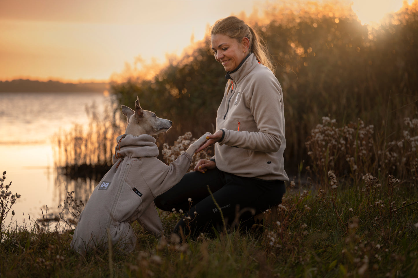 Woman and dog sitting by a lake at sunset, wearing matching colour fleeces.