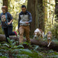 Lifestyle image of the Crag EX Lead, being worn around the neck of a lady, while a man walks beside her with a lead in his hand. Two dogs step over the log behind them.