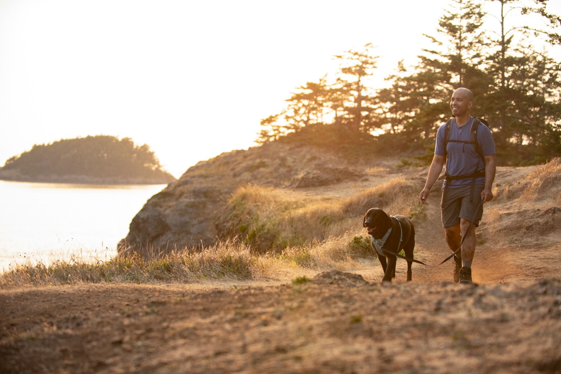 Lifestyle image of the Front Range Lead, clipped into a dog's harness while the dog and their owner walk over some dry terrain, with a body of water and trees in the background.