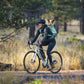 Lifestyle image of the Hitch Hiker Carrier, showing someone riding a bicycle with a dog on their back, surrounded by trees and brush.