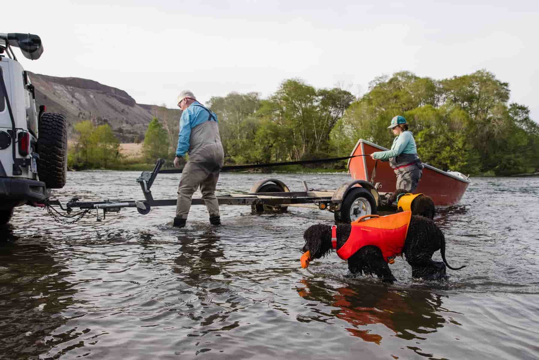 Dog in a body of water, wearing a lifejacket. Two owners are stood behind them near a boat.